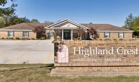 Exterior view of Highland Crest assisted living facility by Americare, showing a single-story brick building with a central entrance and a large sign in front displaying the facility's name.