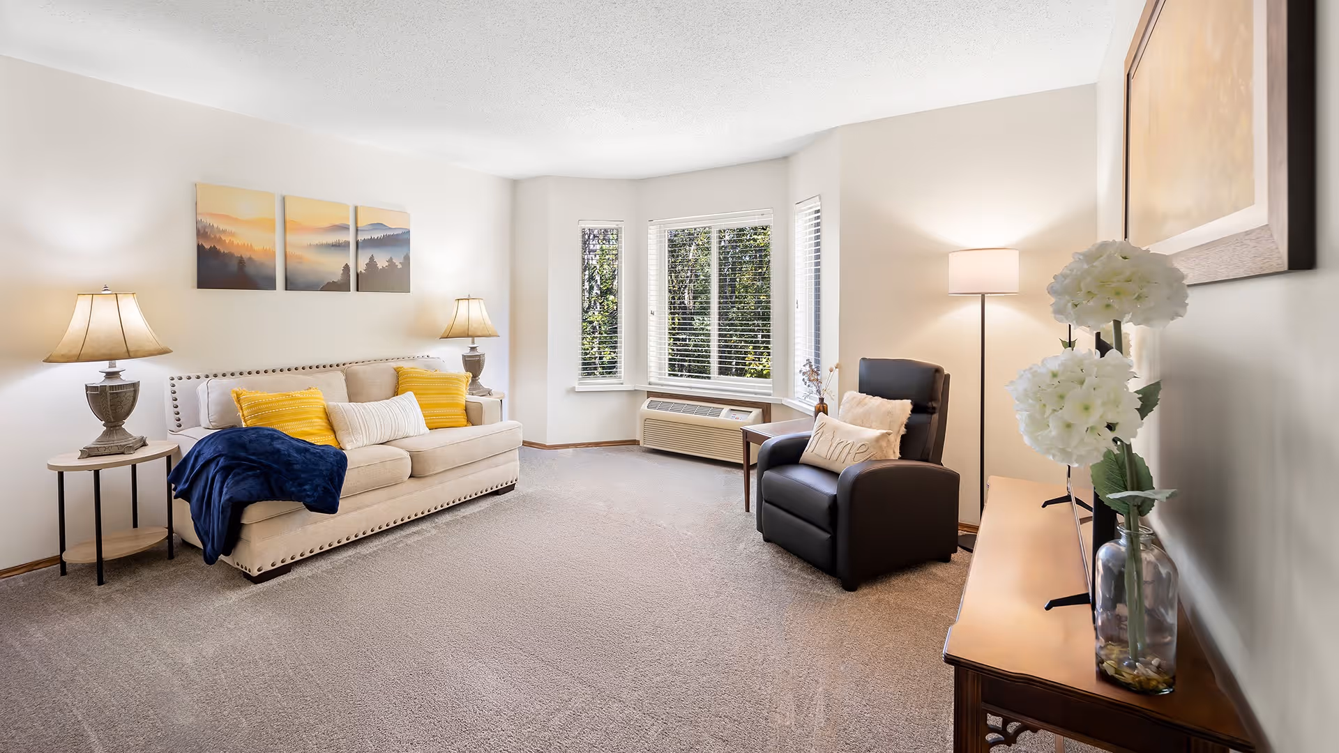 A bright and cozy living room with a beige sofa adorned with yellow and white pillows, a dark blue throw blanket, and two table lamps on either side. There is a black recliner chair with a beige pillow next to a floor lamp. A wooden console table with a vase of white flowers is against the wall, and three-panel artwork depicting a misty forest hangs above the sofa. Large windows with white blinds let in natural light, and the room is carpeted in a neutral tone.