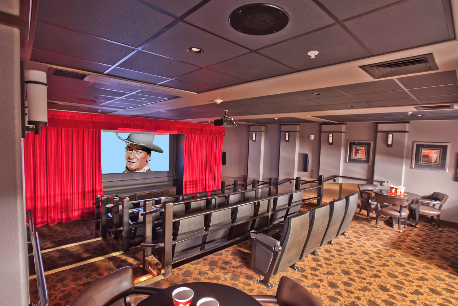 Interior view of a small movie theater room with rows of theater seats facing a screen displaying a man wearing a cowboy hat. The room has red curtains around the screen, patterned carpet, and a few tables with chairs and popcorn cups on the right side.