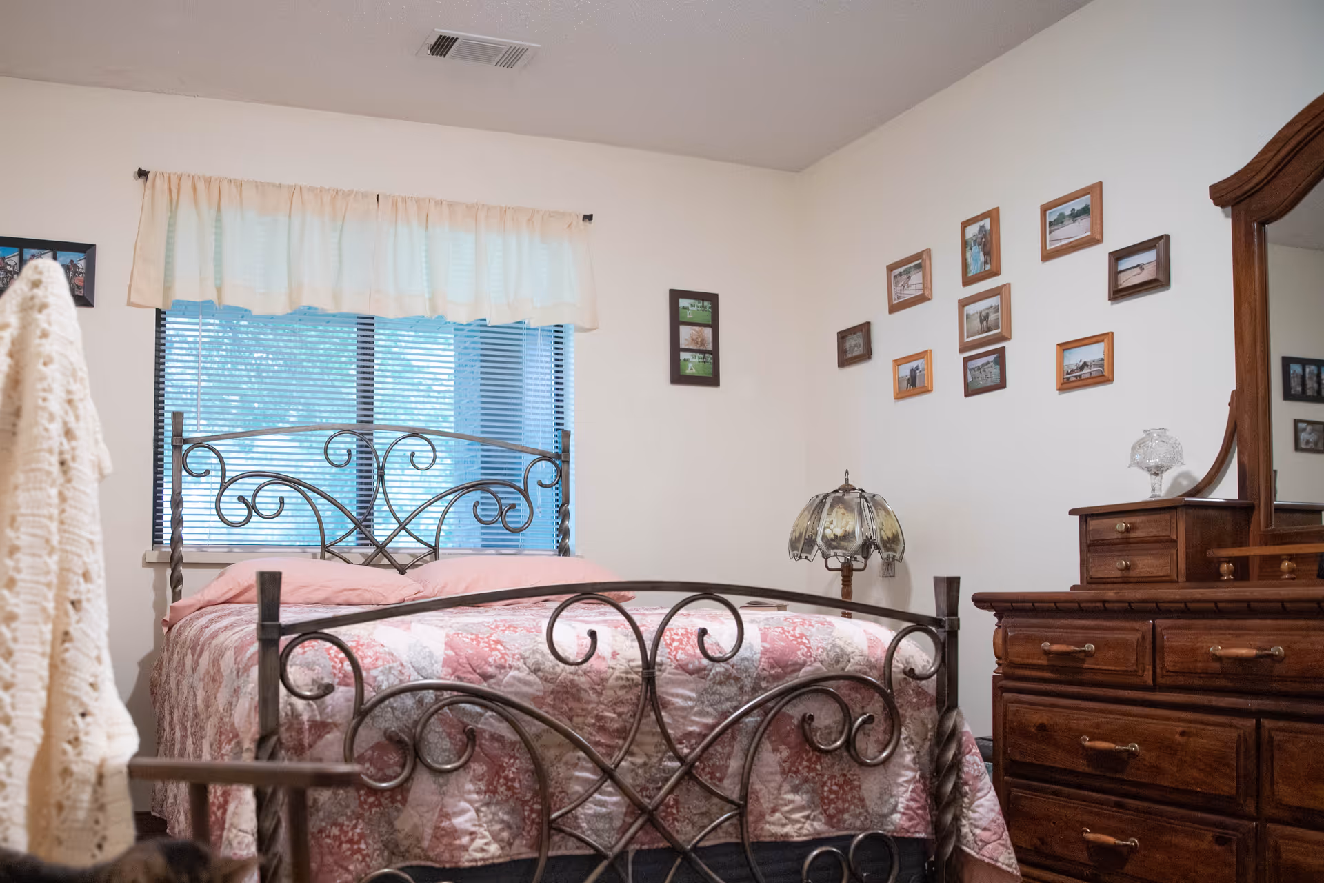 A cozy bedroom with a decorative metal bed frame and a pink patterned quilt. There is a window with blinds and a light-colored valance above the bed. On the right wall, there are multiple framed photos arranged in a grid pattern. A wooden dresser with a mirror and a vintage-style lamp are also visible.