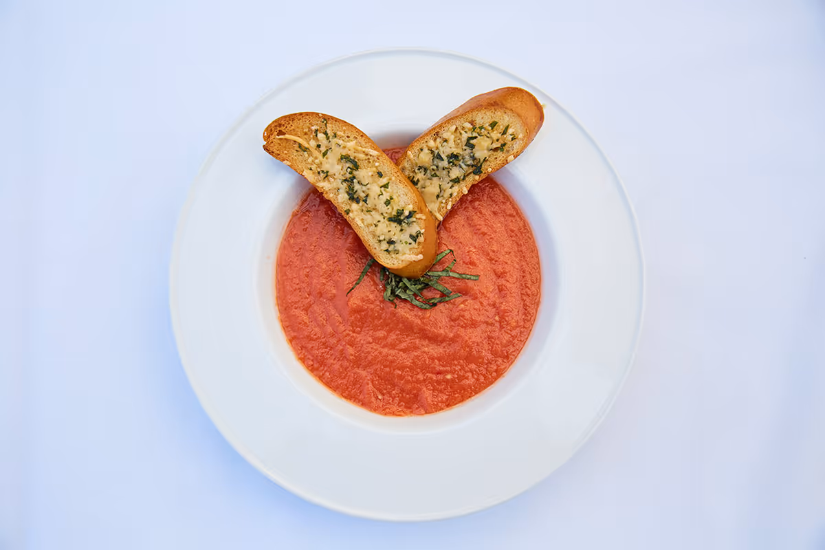 Top-down view of a bowl of tomato soup garnished with basil and two slices of garlic bread on a white plate.