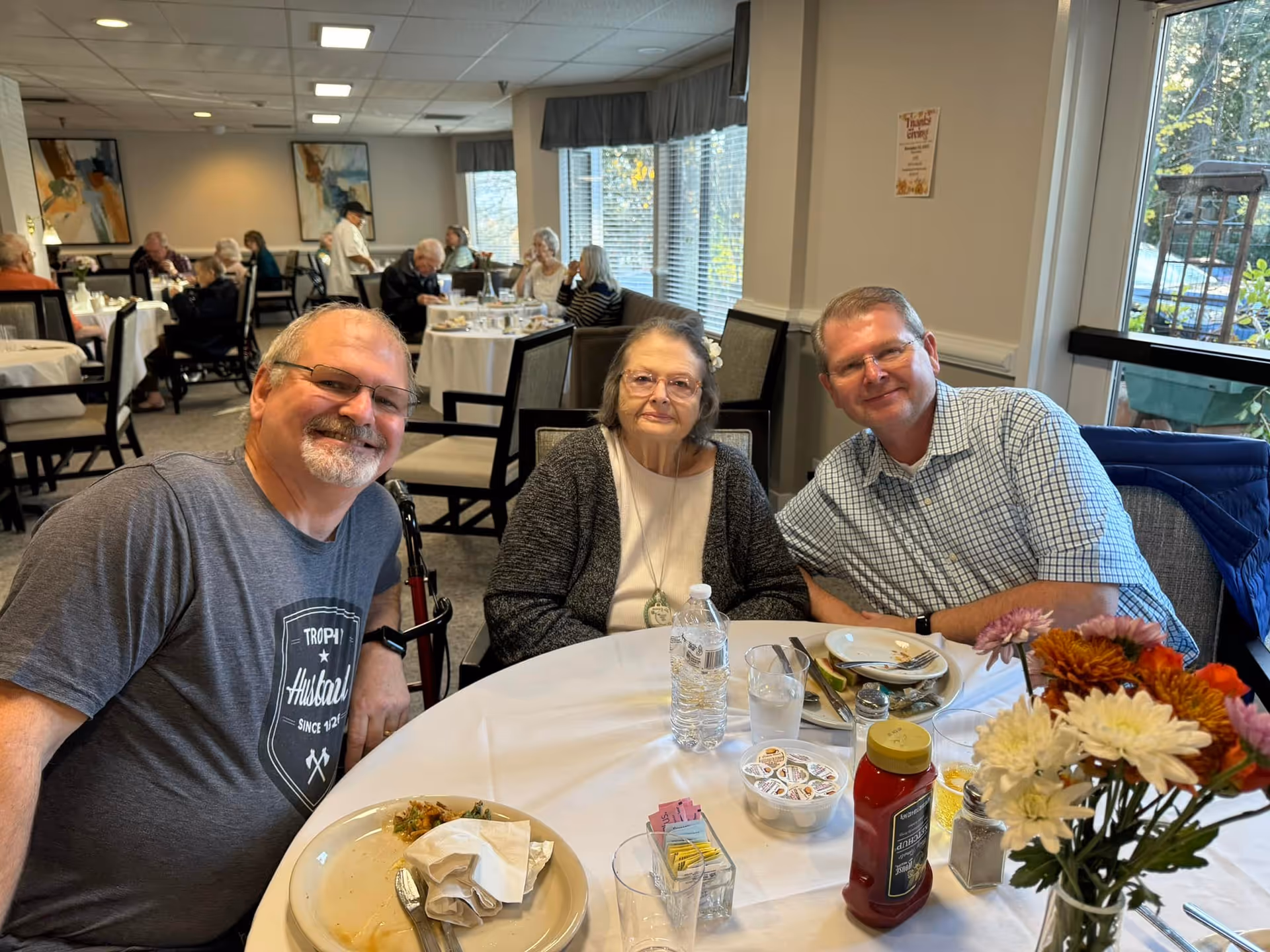 Three people sitting at a round dining table in a senior living facility dining room. Two men and one elderly woman are smiling at the camera. The table has plates, glasses, a bottle of ketchup, a water bottle, and a vase with colorful flowers. Other residents are visible dining in the background near large windows.