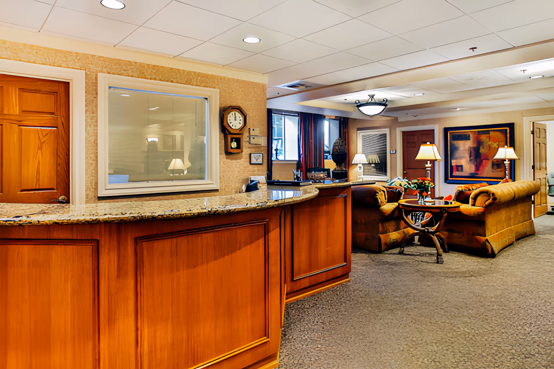 Interior view of a senior living facility reception area with a wooden front desk featuring a granite countertop. Behind the desk is a window and a wall clock. In the background, there is a cozy seating area with upholstered sofas, a round wooden coffee table with a flower vase, several table lamps, and framed artwork on the walls. The space is warmly lit and carpeted.