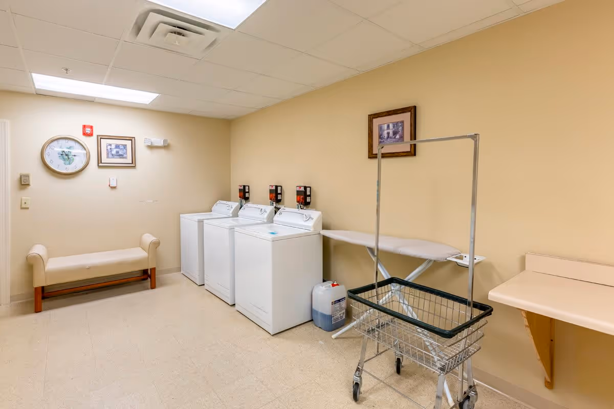 A laundry room with three white washing machines lined up against a beige wall. There is an ironing board and a laundry cart with a metal frame and wheels in front of the machines. On the left side, there is a small beige bench beneath a clock and two framed pictures on the wall. The floor is light-colored tile, and the ceiling has fluorescent lighting.