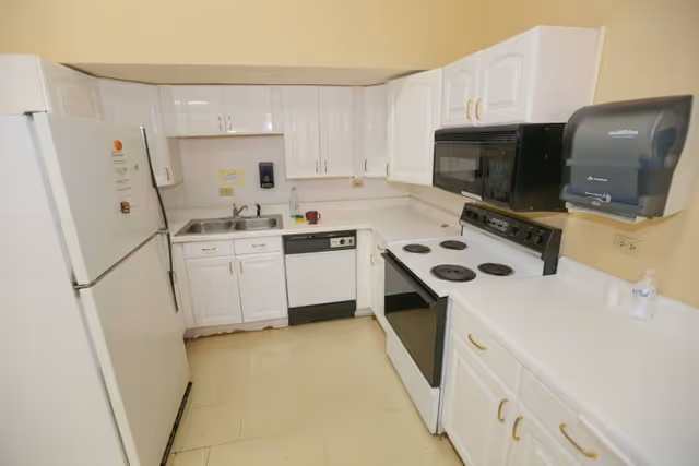 A small kitchen with white cabinets, a white refrigerator, a white electric stove with four burners, a black microwave above the stove, a dishwasher, a double sink, and a wall-mounted paper towel dispenser. The walls are painted light yellow and the floor has beige tiles.