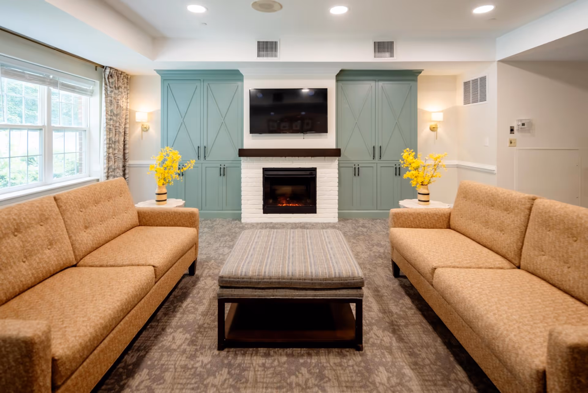 Communal living room with two tan sofas facing a central ottoman, a fireplace beneath a wall-mounted TV flanked by green cabinets and yellow floral arrangements.