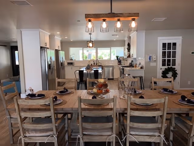 A spacious dining area with a long wooden table set for eight people, each place setting featuring a plate, napkin, and cutlery. Above the table hangs a rustic light fixture with five exposed bulbs. In the background, there is a modern kitchen with white cabinets, stainless steel appliances including a refrigerator and dishwasher, and a kitchen island with bar stools. The room has neutral-colored walls and large windows letting in natural light.