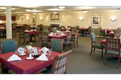 A spacious dining room with multiple tables covered in burgundy tablecloths, each set with white napkins, cups, and saucers. The chairs have green and plaid upholstery. The walls are decorated with framed artwork and sconces providing warm lighting.