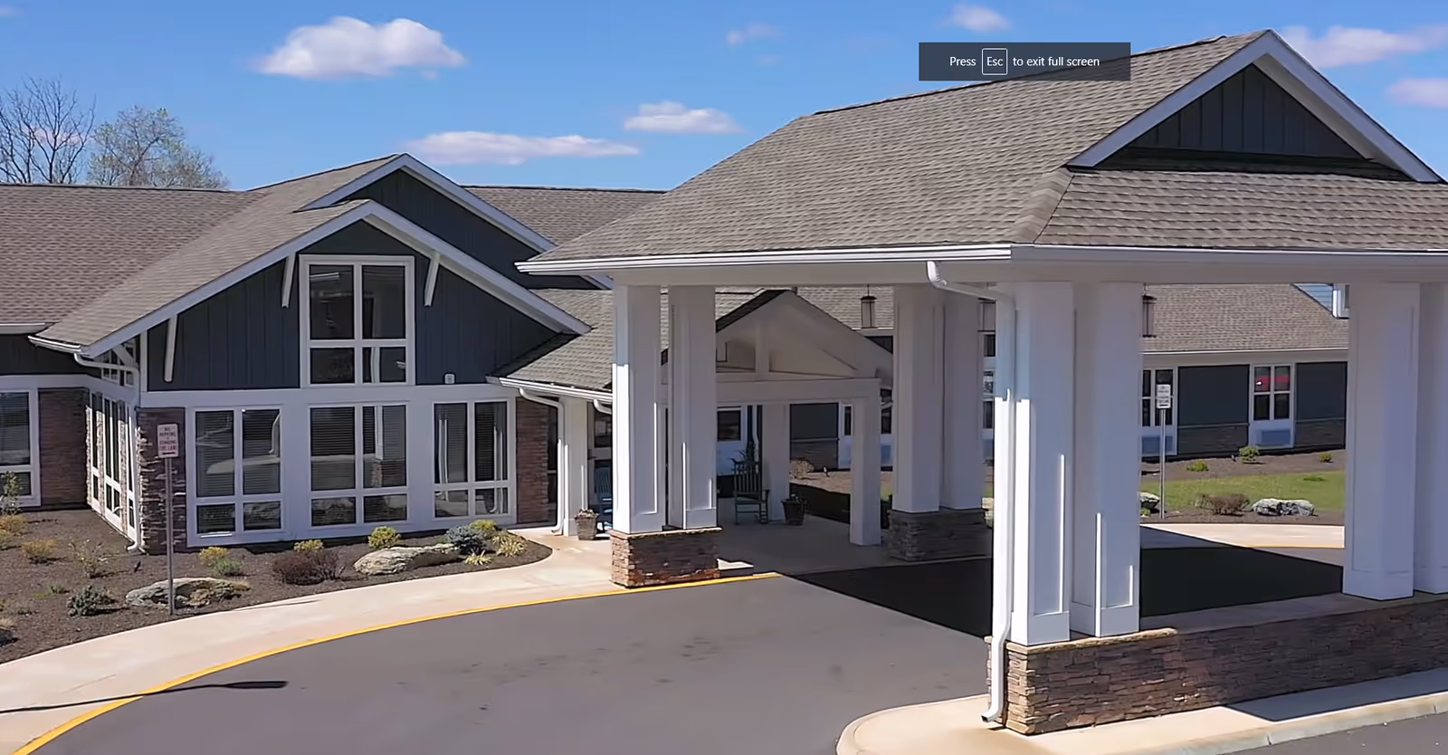 Exterior view of a senior living facility building with a covered entrance supported by white columns and stone accents. The building has large windows and a sloped roof under a clear blue sky.