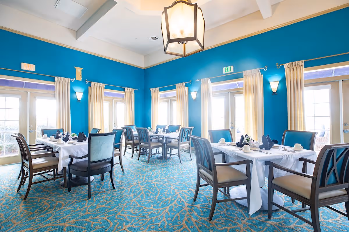 Bright dining room with turquoise walls and patterned carpet, featuring multiple tables set with white tablecloths, napkins, cups, and silverware. The room has large windows with cream-colored curtains allowing natural light to fill the space, and a large hanging light fixture in the center.