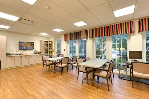 A bright room with wooden flooring featuring two rectangular tables surrounded by chairs. Large windows with striped valances let in natural light and offer a view of greenery outside. Cabinets and shelves line one wall, with a flat-screen TV mounted above them. A computer workstation with a chair is positioned near the windows.