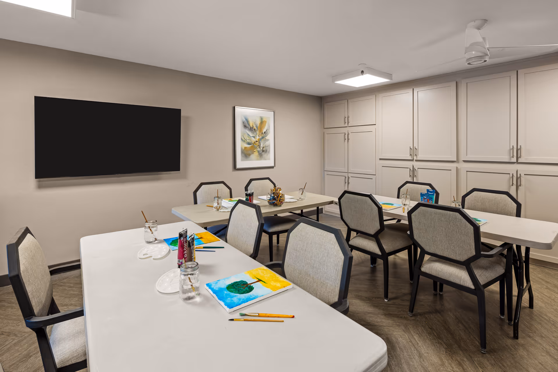 A well-lit room with several tables and chairs arranged for an art activity. On the tables are painting supplies including brushes, paint tubes, jars of water, and paintings in progress. The room has beige walls, a large flat-screen TV mounted on one wall, a framed abstract painting, and built-in cabinets along another wall.