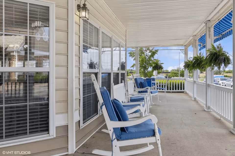 Covered porch area with white rocking chairs featuring blue cushions lined up against the beige siding of a building. The porch has white railings and hanging green plants, with a view of trees and parked cars in the background under a blue sky.