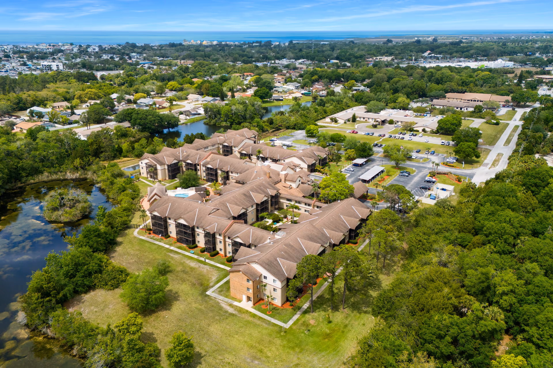 Aerial view of a large senior living facility complex surrounded by greenery, trees, and small bodies of water. The facility has multiple connected buildings with brown roofs and parking lots. Residential neighborhoods and a distant coastline are visible in the background under a clear blue sky.
