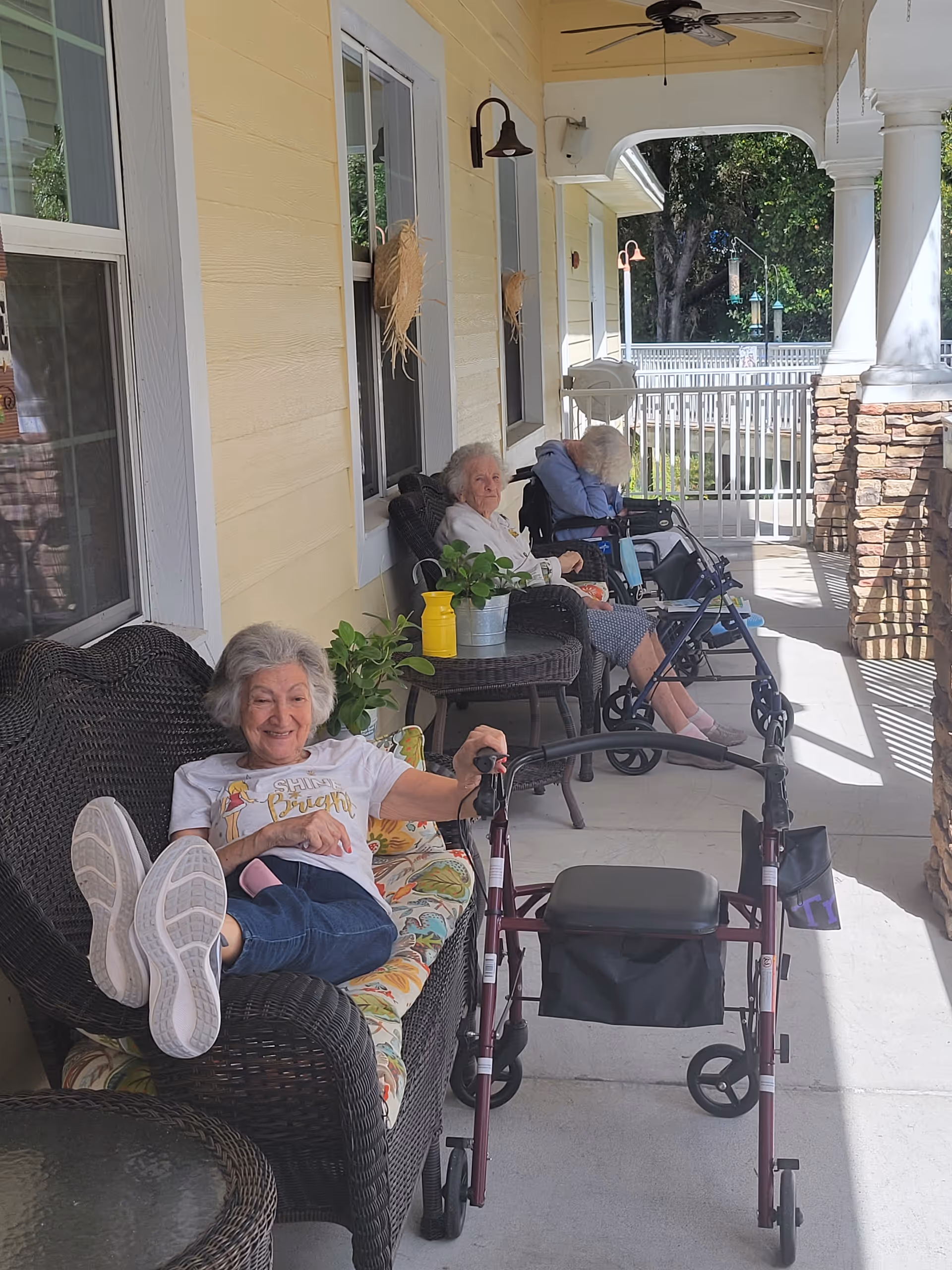 Three older adults relaxing on a covered porch with wicker chairs, plants, and walkers at an assisted living facility.