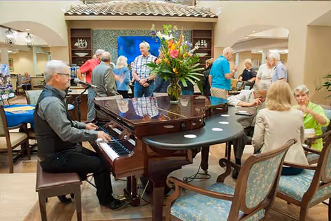 A man playing a grand piano in a common area of a senior living facility with several elderly people standing and sitting around, engaging in conversation. The room has comfortable chairs, a large floral arrangement on the piano, and warm lighting.
