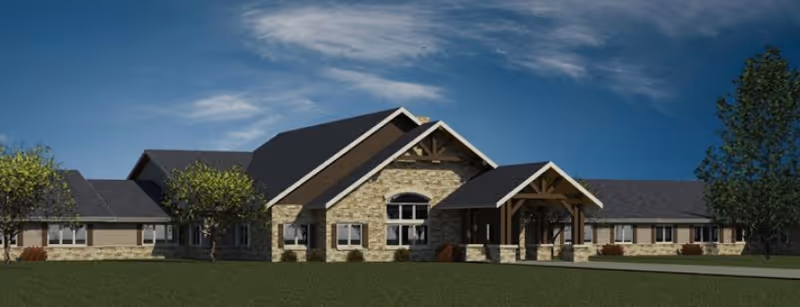 Exterior view of a single-story retirement living facility with stone and wood siding, multiple windows, and a covered entrance under a peaked roof, set against a blue sky with some clouds and surrounded by green grass and a few trees.