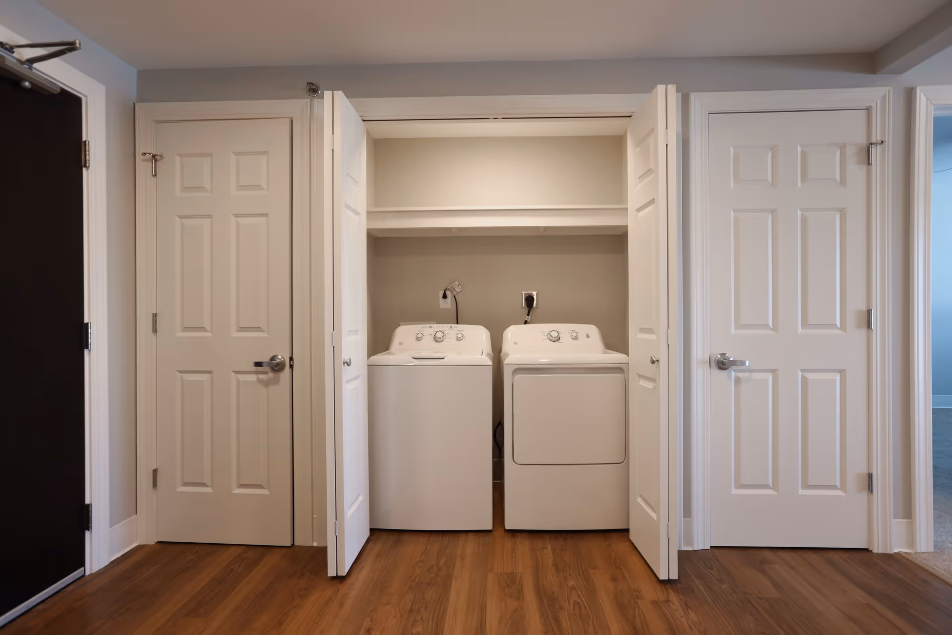 A laundry area with a white washing machine and dryer placed side by side inside a closet with bi-fold doors open. The floor is wooden, and there are two closed white doors on either side of the laundry closet.