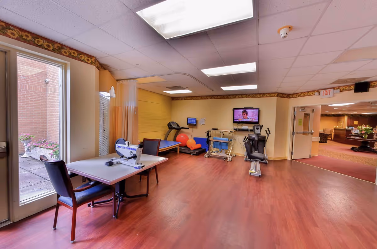 A rehabilitation or physical therapy room with exercise equipment including a treadmill, exercise bike, therapy table, and therapy balls. There is a table with a chair and a small pedal exerciser on it near a glass door leading outside. The room has wood flooring, yellow walls with sunflower border trim, and ceiling lights. A TV is mounted on the wall showing a program. An open door leads to a carpeted common area with seating and a reception desk.