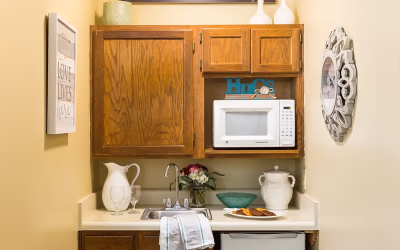 Small kitchenette area with wooden cabinets, a white microwave, a sink with a faucet, a white pitcher, two wine glasses, a vase with flowers, a green bowl, a plate with cookies, and a decorative wall clock on the right wall.