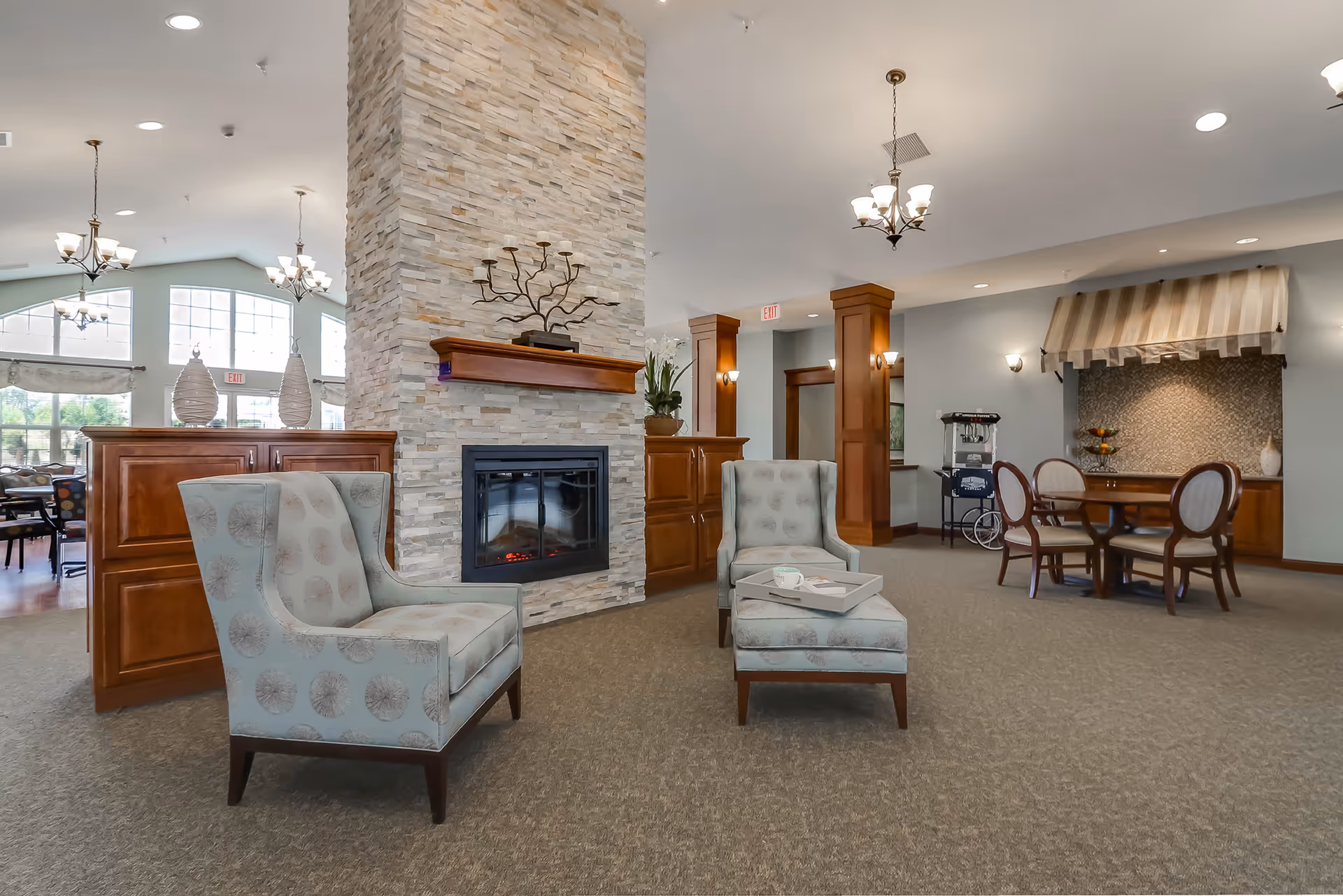 A spacious and well-lit common area in an assisted living facility featuring a stone fireplace with a wooden mantle, two upholstered armchairs with a matching ottoman, a small table with a cup and tray, wooden cabinetry, a dining table with four chairs, and large windows allowing natural light to fill the room.