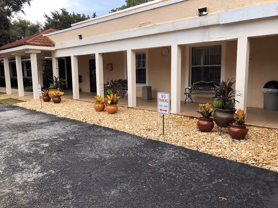 Exterior view of a building with a covered walkway supported by white columns. Several potted plants are arranged along a gravel bed next to a paved area. There are benches under the walkway and a 'No Parking Fire Lane' sign is visible in the gravel area.