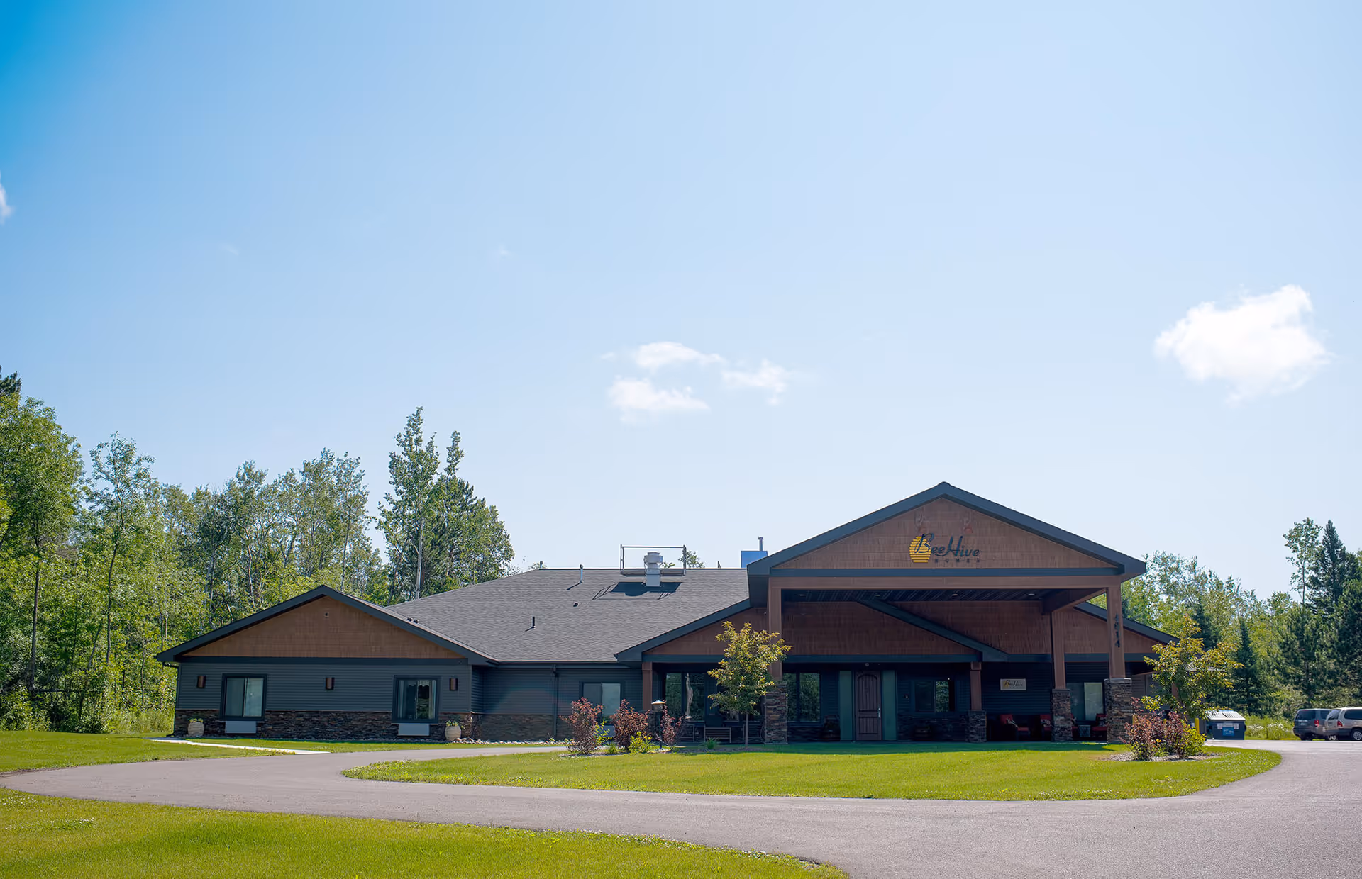 Exterior view of BeeHive Homes Assisted Living and Memory Care of Duluth, a single-story building with a covered entrance, surrounded by green grass and trees under a clear blue sky.