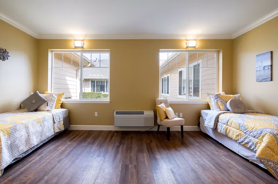 A bright and clean bedroom with two single beds on opposite sides of the room, each with yellow and gray bedding and multiple pillows. Between the beds are two large windows with views of the exterior building and a beige upholstered chair with pillows. The room has wooden flooring and light yellow walls with white trim and crown molding. A small framed picture hangs on the right wall.
