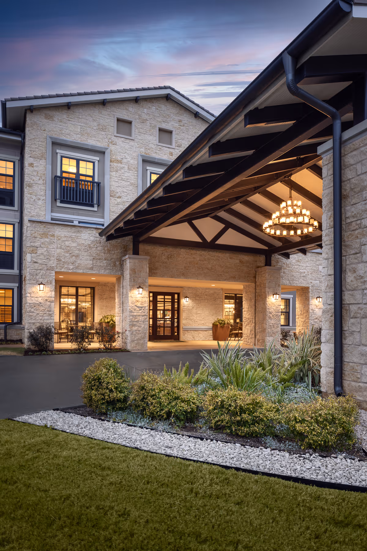 Entrance of Belmont Village Senior Living Lakeway at dusk, featuring a stone building with large windows, a covered driveway with wooden beams, a chandelier hanging from the ceiling, and landscaped greenery with bushes and plants in the foreground.