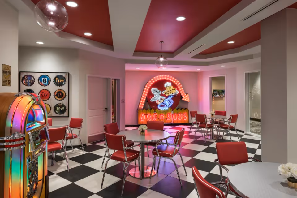 A retro-themed dining area with black and white checkered floor tiles, red chairs, and round tables. A colorful jukebox is on the left side, and a neon sign reading 'DOG n SUDS' with a cartoon dog holding a tray of food is displayed on the back wall. The ceiling has a red and white design with recessed lighting and hanging glass globe lights. There are framed vintage-style signs on the wall and doorways leading to other rooms.