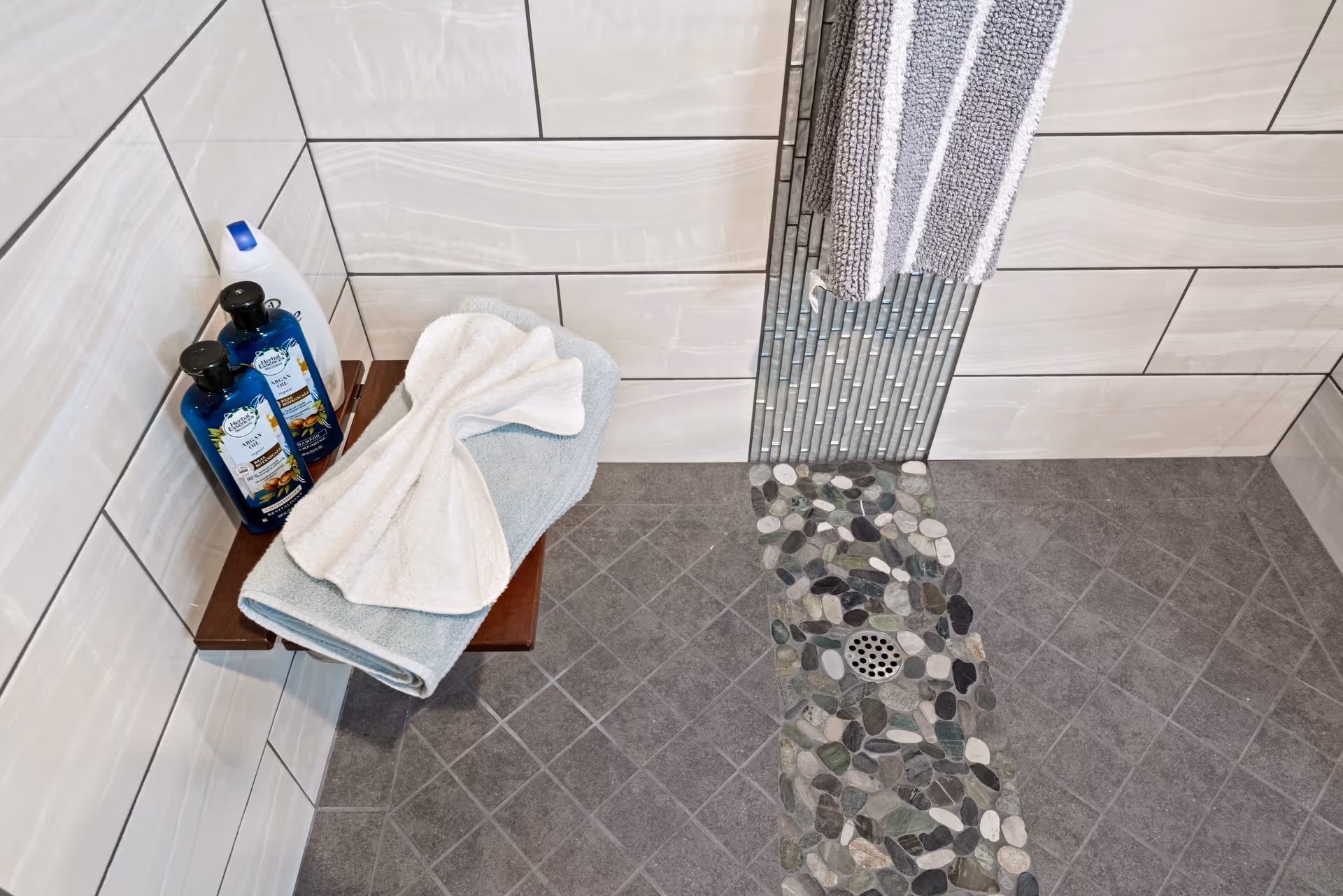 A corner of a shower area with a tiled floor featuring a pebble stone drainage strip, light-colored wall tiles, a wooden shower bench holding folded towels and bottles of shampoo and body wash, and a striped towel hanging on the wall.