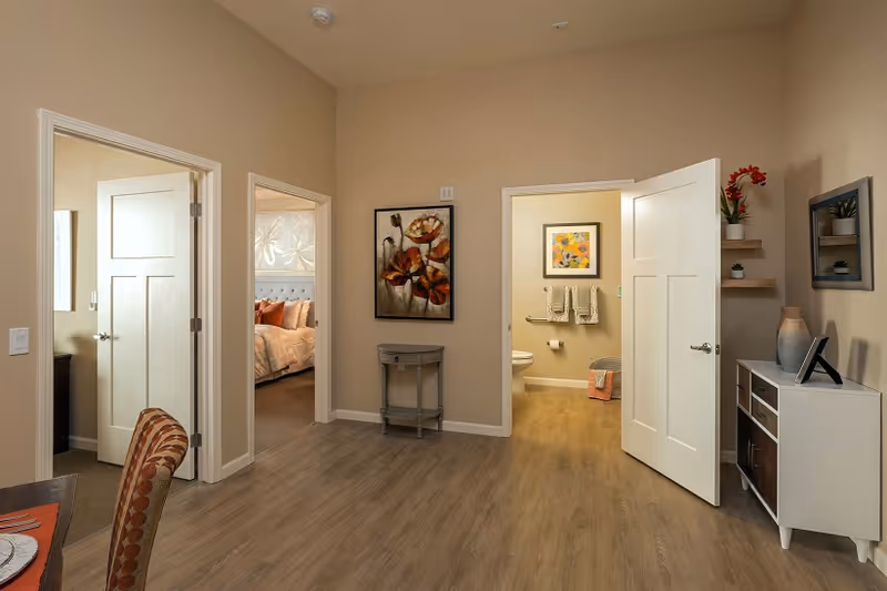 Interior view of a furnished senior living unit showing a central hallway with open doors to a bedroom and bathroom, wood floors, and neutral decor.