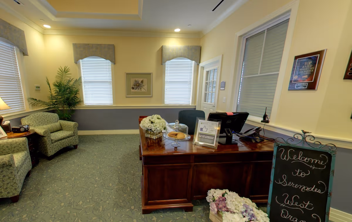 Reception/welcome area of a senior living facility with a wooden front desk, seating chairs, floral arrangements, and a chalkboard welcome sign.