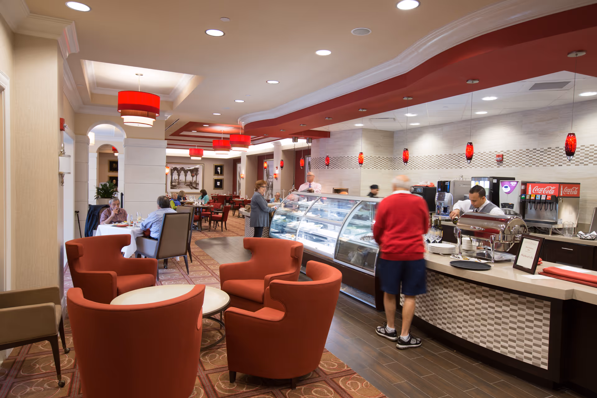 Interior view of a dining area in a senior living facility with red cushioned chairs and round tables in the foreground. People are seated and eating at tables in the background. A curved counter with a glass display case and beverage dispensers is on the right side, with staff serving food. Red pendant lights hang from the ceiling.