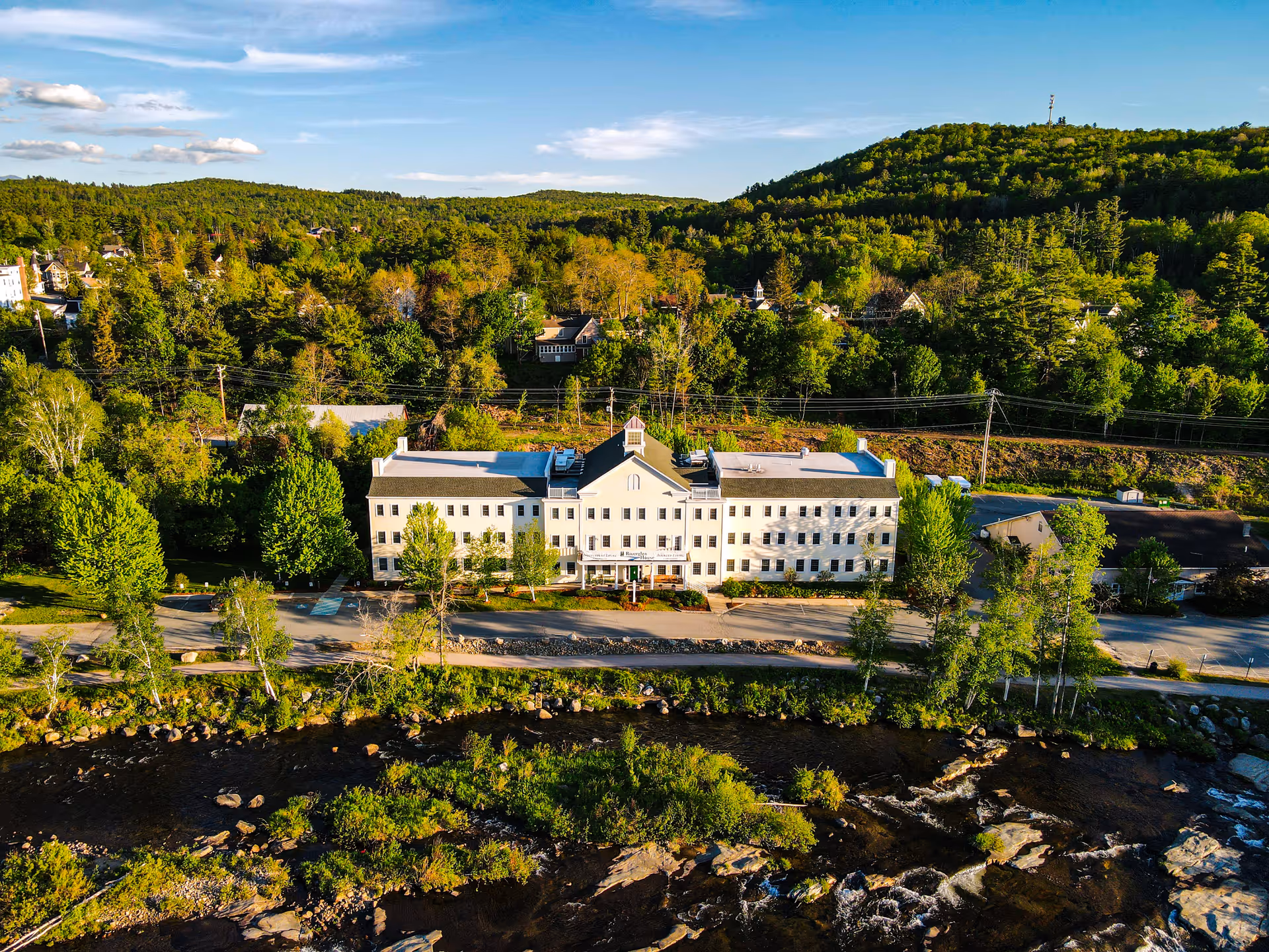 Aerial view of a large, white, three-story building named Riverglen House surrounded by trees and greenery, with a river flowing in the foreground and forested hills in the background under a blue sky with scattered clouds.