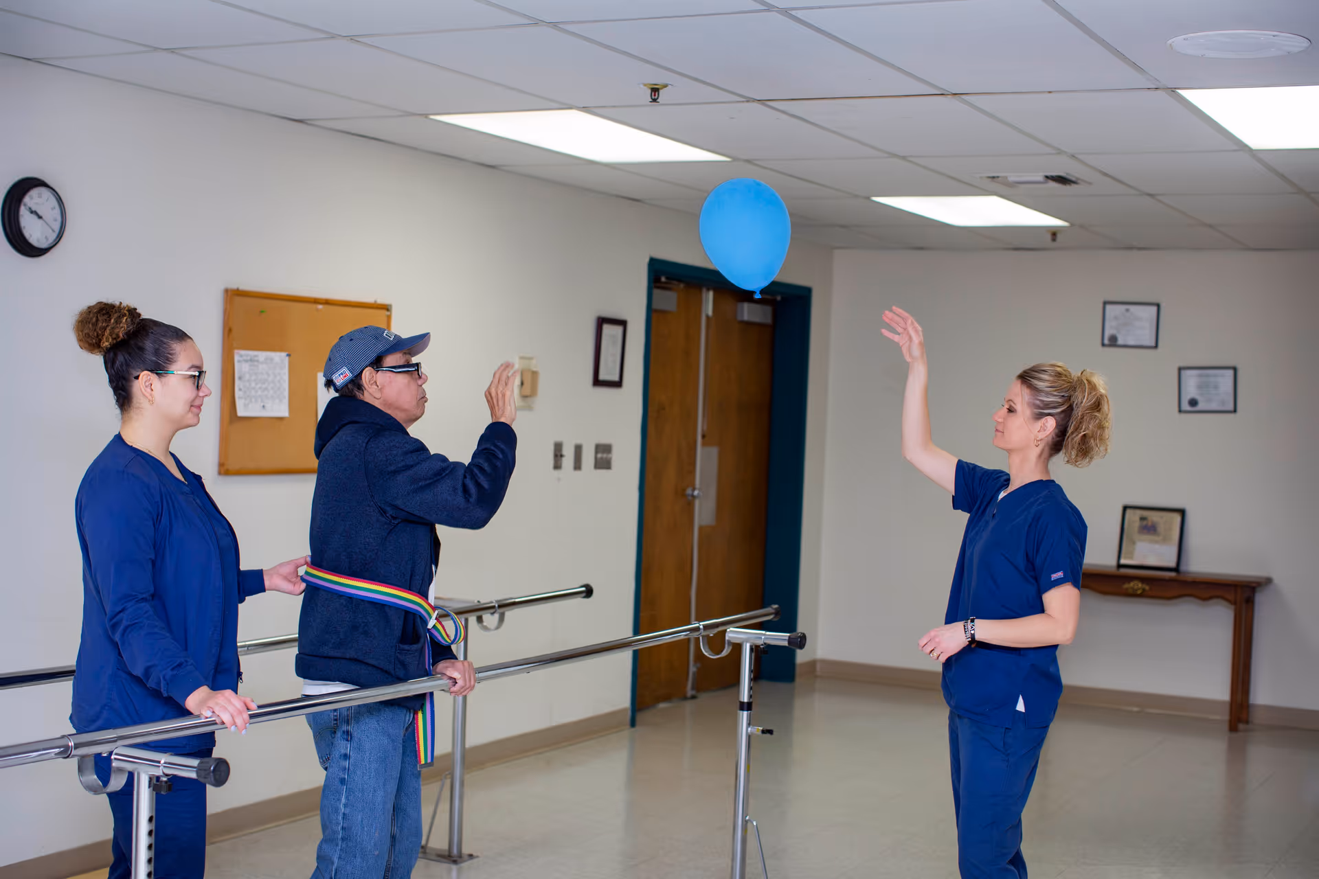 A man wearing a cap and a support belt is practicing raising his hand to hit a blue balloon while standing between parallel bars in a rehabilitation or therapy room. Two female healthcare workers in blue scrubs assist and encourage him. The room has white walls, a clock, a bulletin board, and framed certificates on the wall.