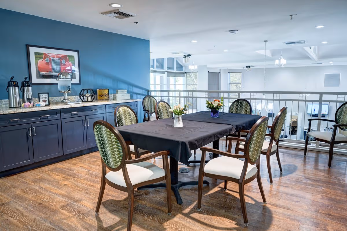 A dining area with two tables covered in black tablecloths, surrounded by chairs with green patterned backs and white seats. On the tables are small vases with flowers. Against the blue wall is a countertop with coffee dispensers, a water dispenser, and decorative items. A framed picture of a red vintage car hangs on the wall above the countertop. The space has wooden flooring and large windows in the background.