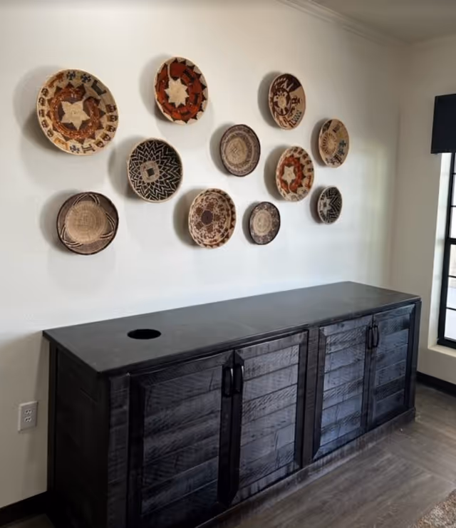 A black wooden cabinet with four doors is placed against a white wall. Above the cabinet, there is a decorative arrangement of eleven woven baskets with various patterns and colors mounted on the wall. To the right, there is a window with black framing allowing natural light into the room.