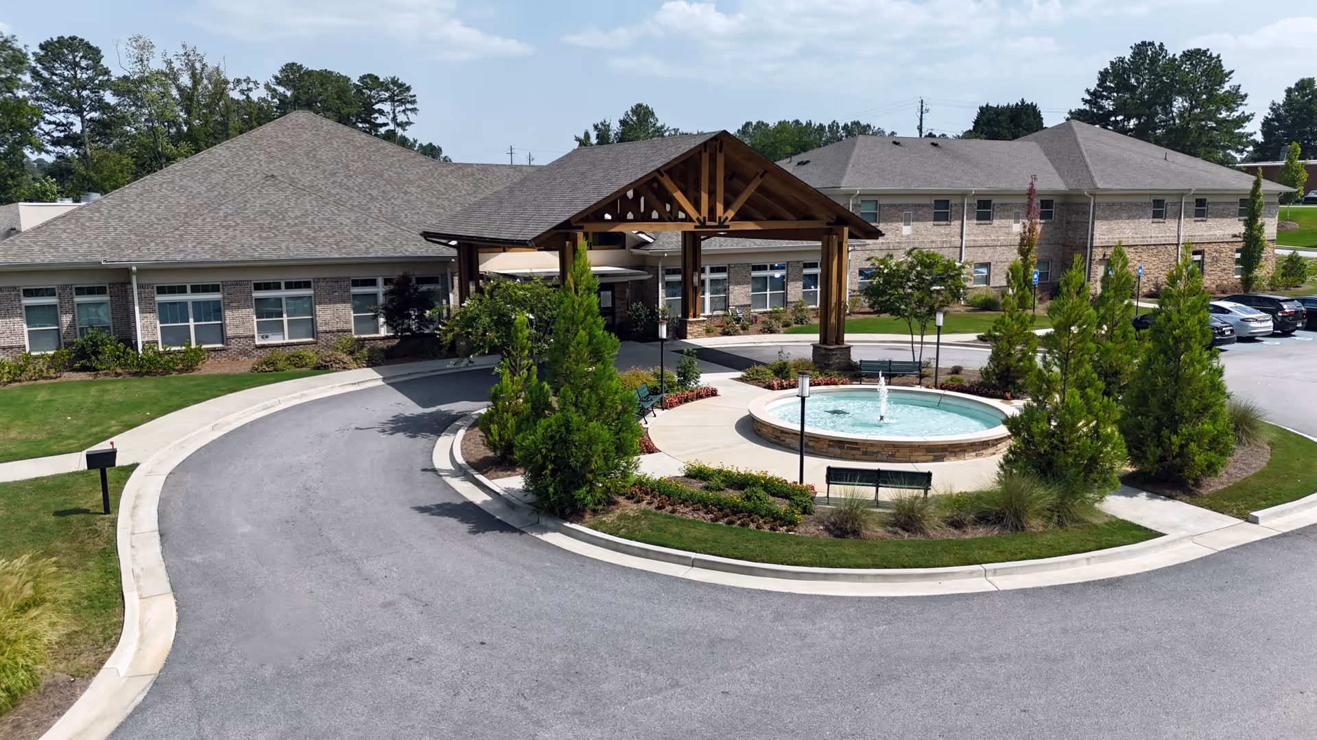 Exterior view of a senior living facility with a circular driveway, a covered entrance with wooden beams, a round fountain surrounded by greenery and benches, and a parking area with cars. The building has brick walls and multiple windows.