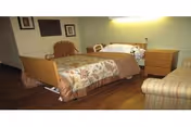 A nursing home bedroom with a single bed covered with a floral patterned blanket and brown bedspread. Next to the bed is a wooden nightstand with a lamp. There is a cushioned armchair with striped upholstery and a wooden chair in the corner. The floor is wooden, and the walls are light-colored with framed pictures hanging.