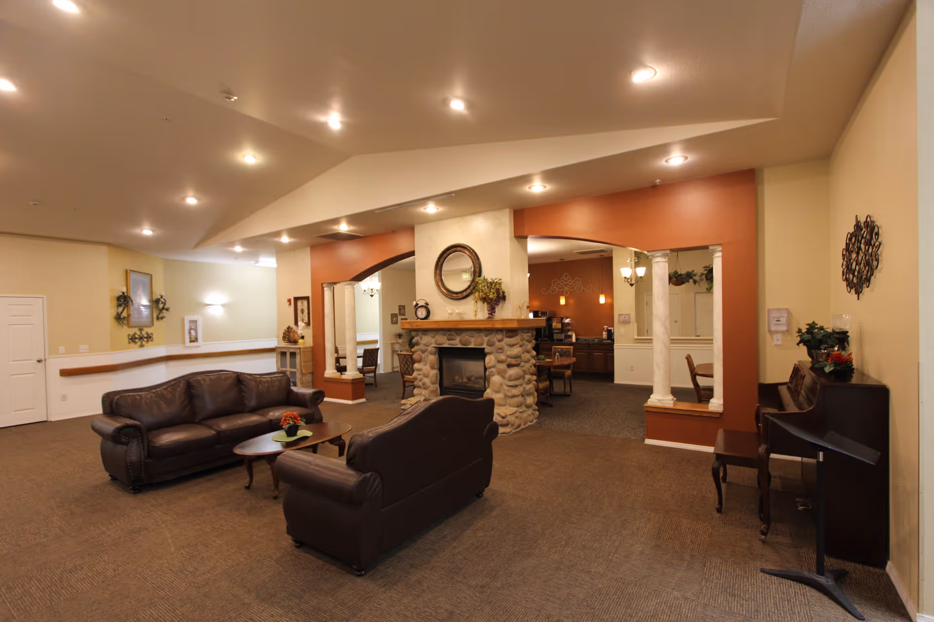A spacious living room area in a retirement facility featuring two dark brown leather sofas arranged around a small wooden coffee table with a flower centerpiece. The room has a stone fireplace with a round mirror and decorative items on the mantel. The walls are painted in warm tones with recessed lighting in the ceiling. In the background, there is a dining area with tables and chairs, and a piano with a music stand is positioned against the right wall.