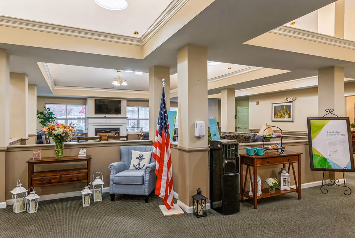 Interior view of a senior living facility common area with beige walls and carpeted floor. There is a blue armchair with an anchor pillow, an American flag, a wooden table with flowers and a phone, a water dispenser, and a small table with mugs and snacks. In the background, there is a fireplace with a TV mounted above it and windows letting in natural light.