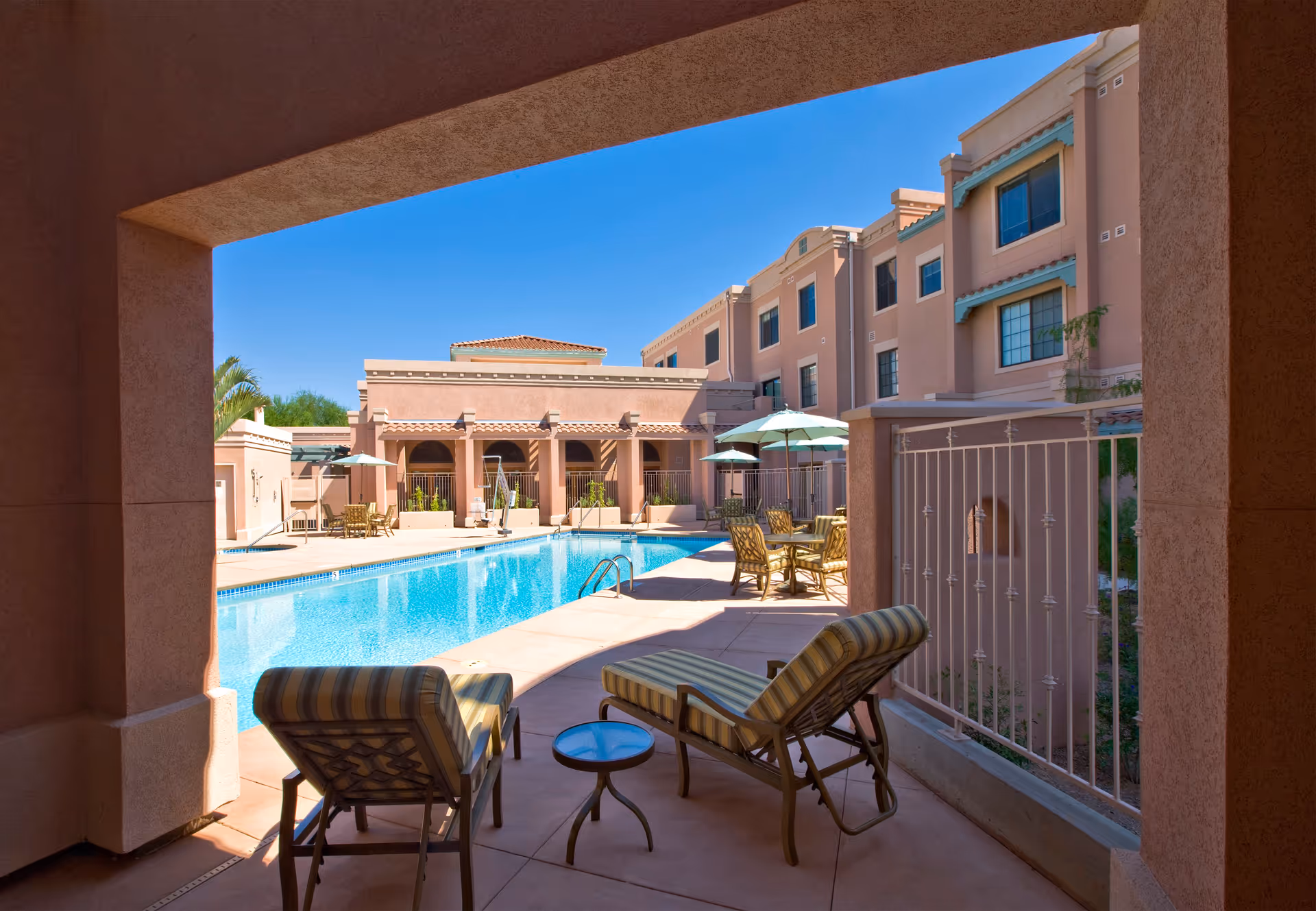View of an outdoor swimming pool area at a senior living facility with lounge chairs and tables with umbrellas around the pool. The building has a southwestern architectural style with stucco walls and multiple windows under a clear blue sky.