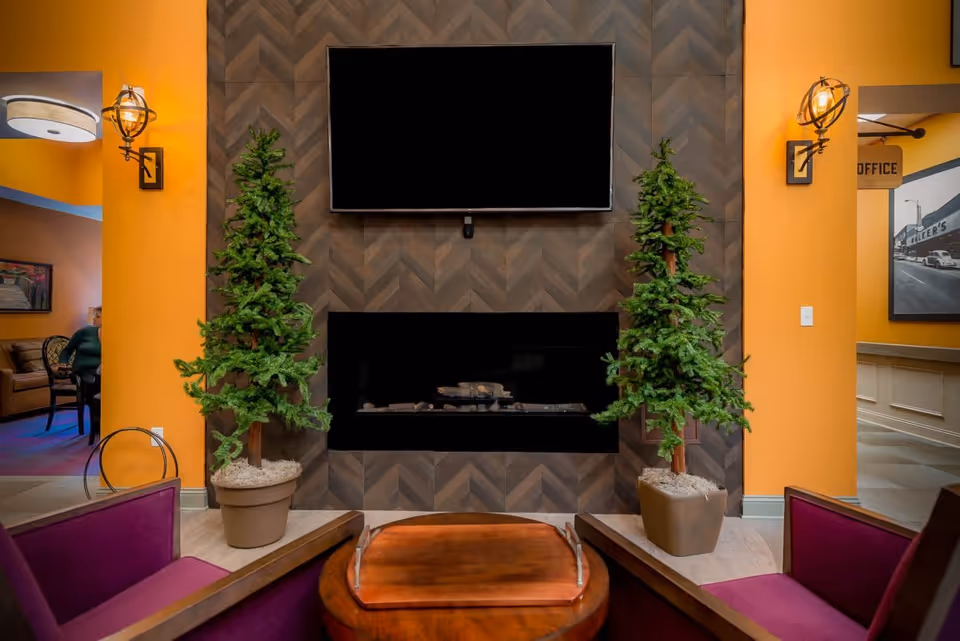 A cozy living room area with two potted artificial evergreen trees flanking a modern fireplace set in a dark herringbone patterned wall. Above the fireplace is a large flat-screen TV. The walls are painted a warm orange color. In the foreground, there are two purple cushioned chairs and a wooden coffee table with a serving tray. To the left, a person is seated on a couch in an adjacent room. To the right, a hallway leads to an office area with a sign and framed artwork on the wall.