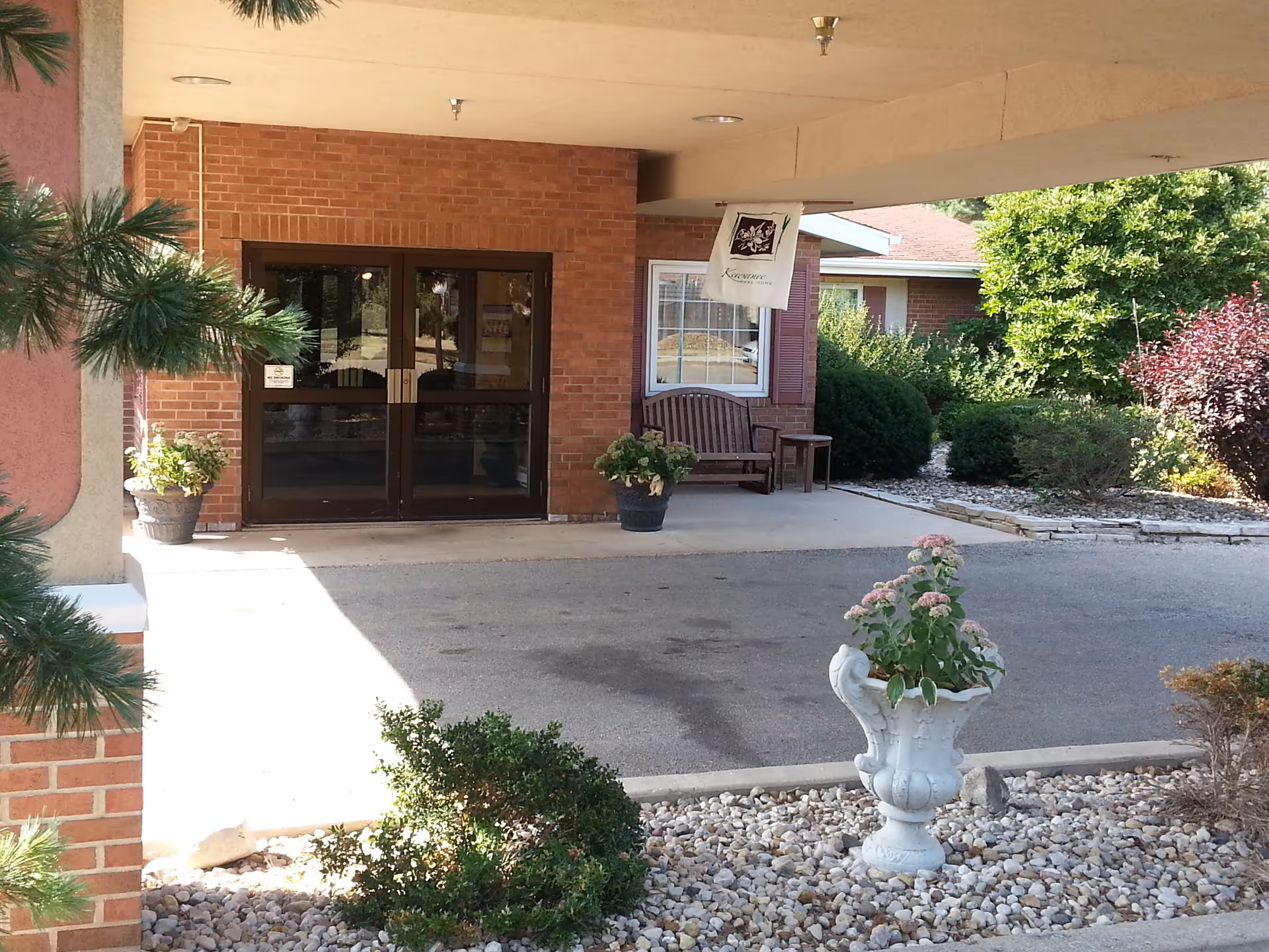 Covered entrance and driveway with double glass doors, a bench, potted plants, and landscaping.