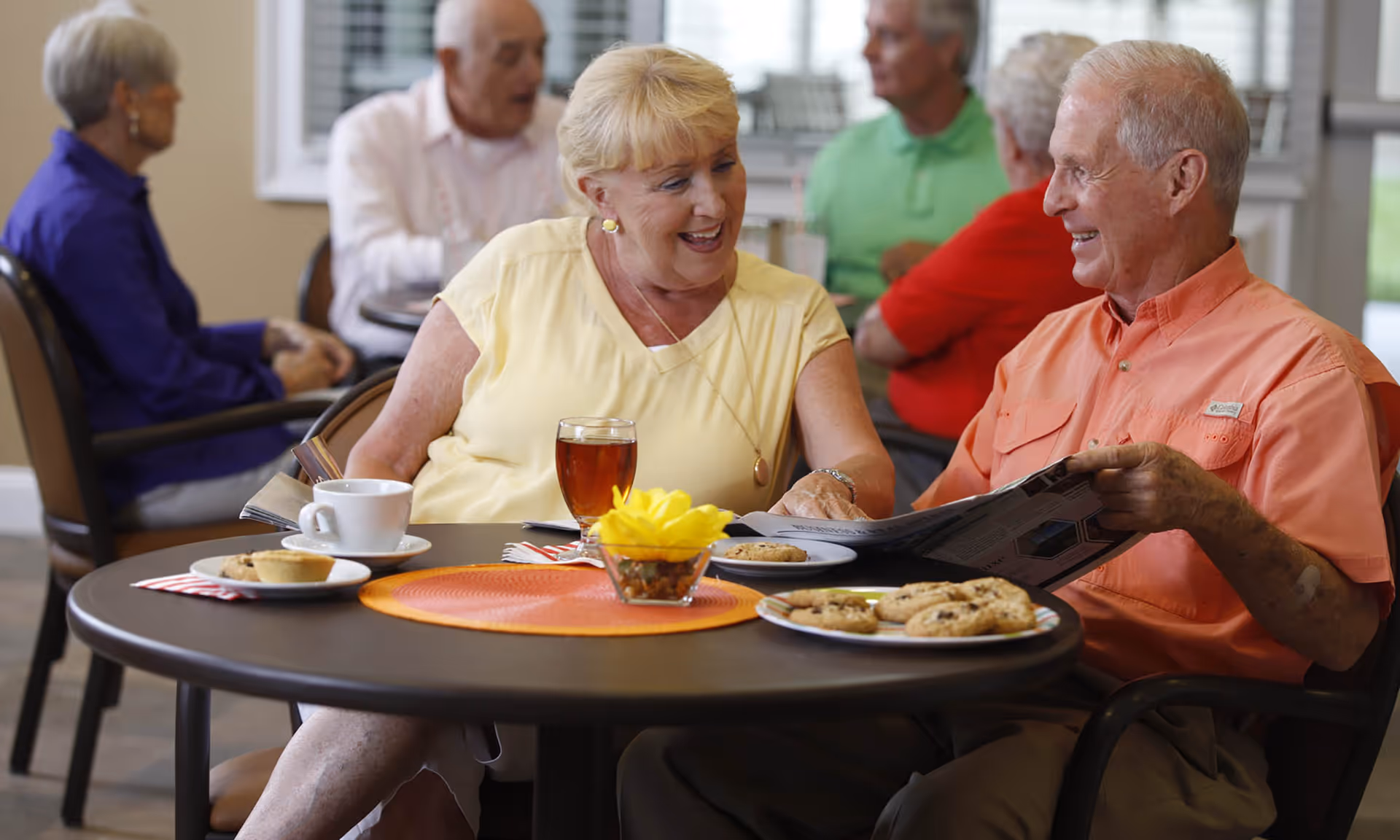 Two elderly people sitting at a round table in a communal dining area, smiling and engaging in conversation. The table has a cup of coffee, a glass of iced tea, a plate of cookies, and a small decorative flower arrangement. In the background, other elderly individuals are seated and conversing.