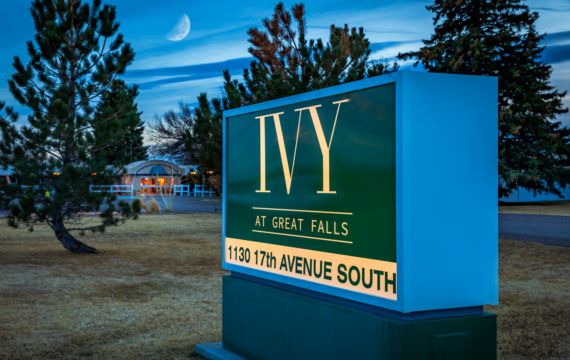 Illuminated green sign reading "IVY AT GREAT FALLS" with the address in front of trees and a distant building at dusk.