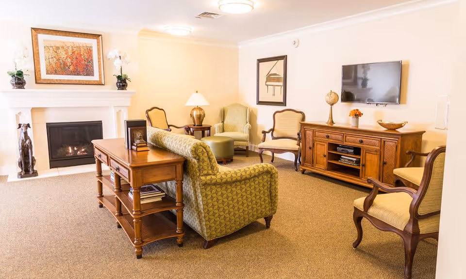 A cozy living room in a senior living facility with a patterned green sofa, wooden side table, several upholstered armchairs, a fireplace with a colorful painting above it, a wooden media console with a flat-screen TV mounted on the wall, and decorative items including a lamp, globe, and flowers.