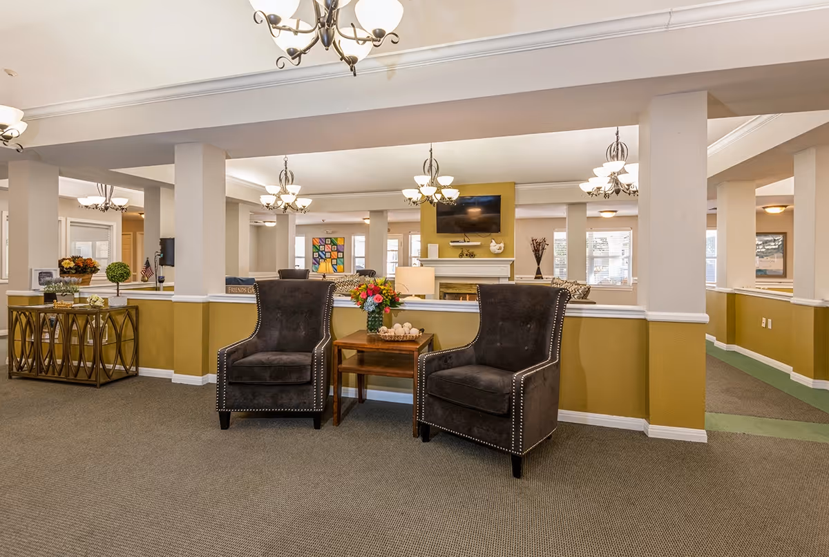 A spacious and well-lit living room area in a senior living facility featuring two dark upholstered armchairs with nailhead trim, a wooden side table with a vase of flowers and decorative items, multiple chandeliers hanging from the ceiling, a fireplace with a mounted flat-screen TV above it, and large windows allowing natural light to fill the room.
