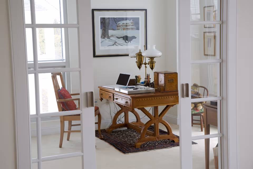 A bright home office seen through glass French doors with a wooden desk, chair, decorative lamp, and framed artwork.